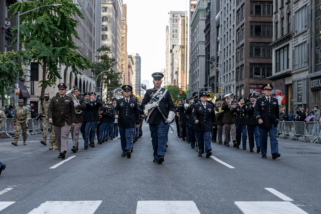 Military parade honoring Veterans Day with uniformed personnel marching in formation along a city street, symbolizing service and sacrifice.