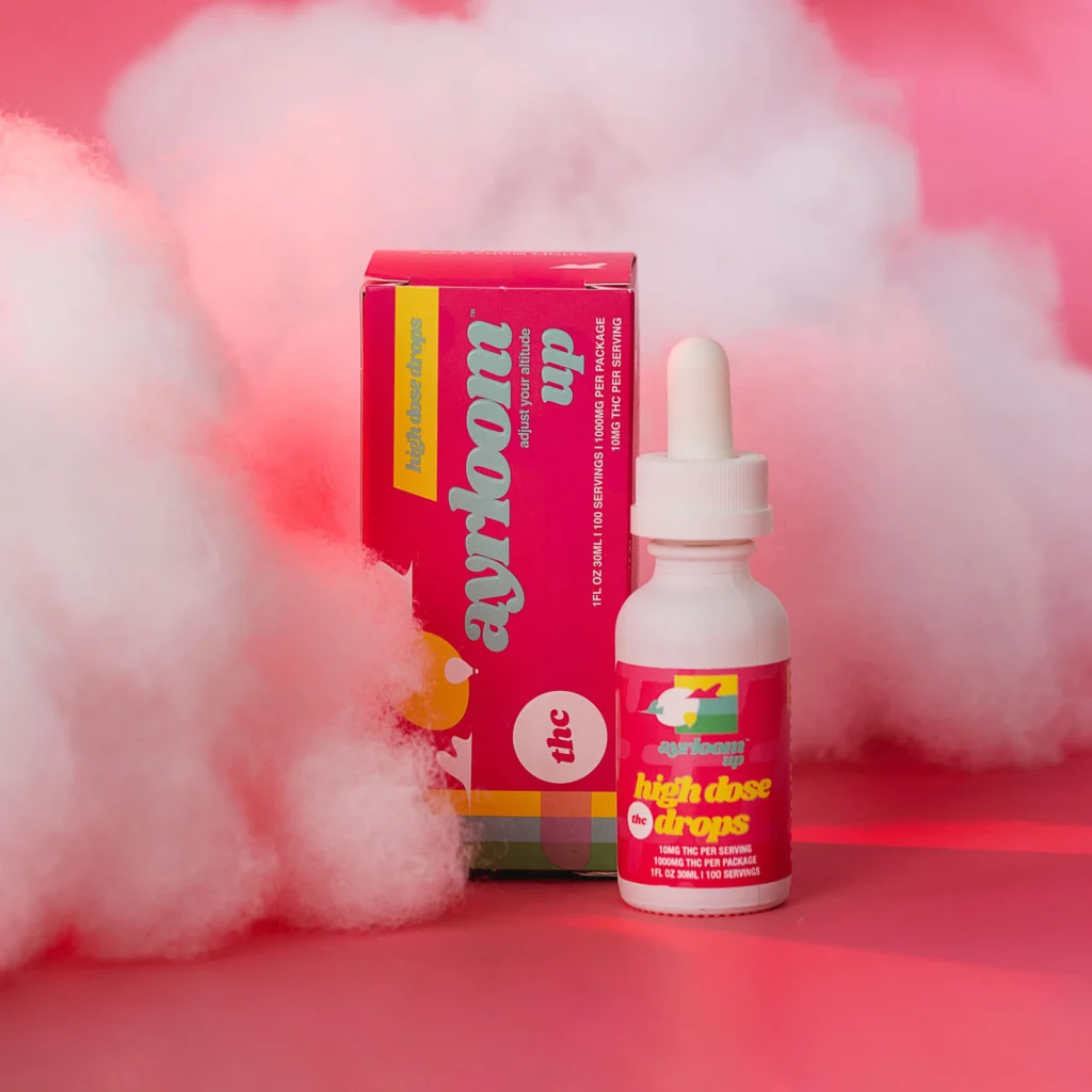 Ayrloom high dose cannabis tincture bottle with colorful packaging, featuring "high dose drops" label, set against a pink background with fluffy white clouds, emphasizing fast-acting cannabis edibles.